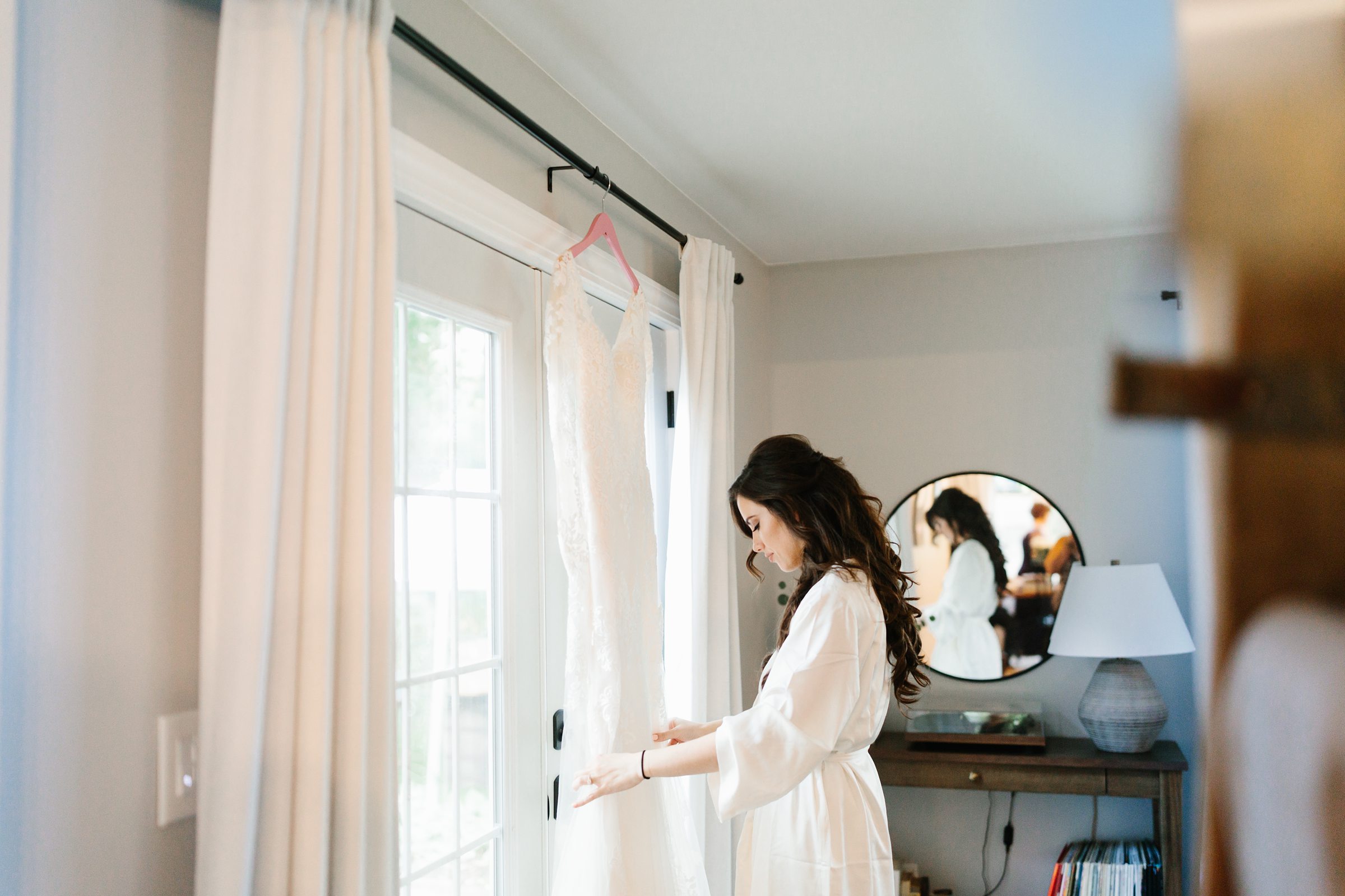 Bride in her robe looks at wedding dress hanging from the window by Detroit Wedding Photographer Michele Maloney 