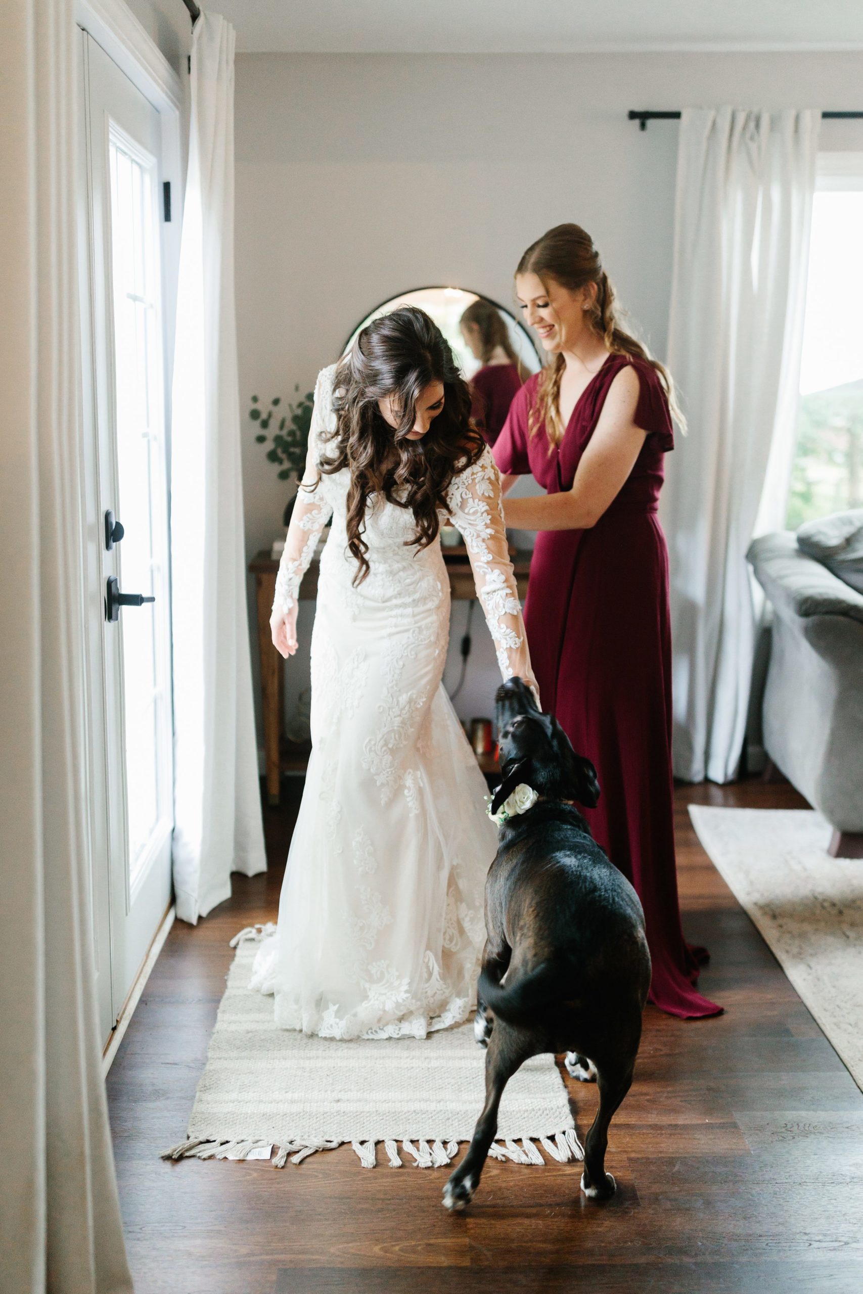 Bridesmaid helps to button up bride's wedding dress while bride pets her dog by Detroit Wedding Photographer Michele Maloney