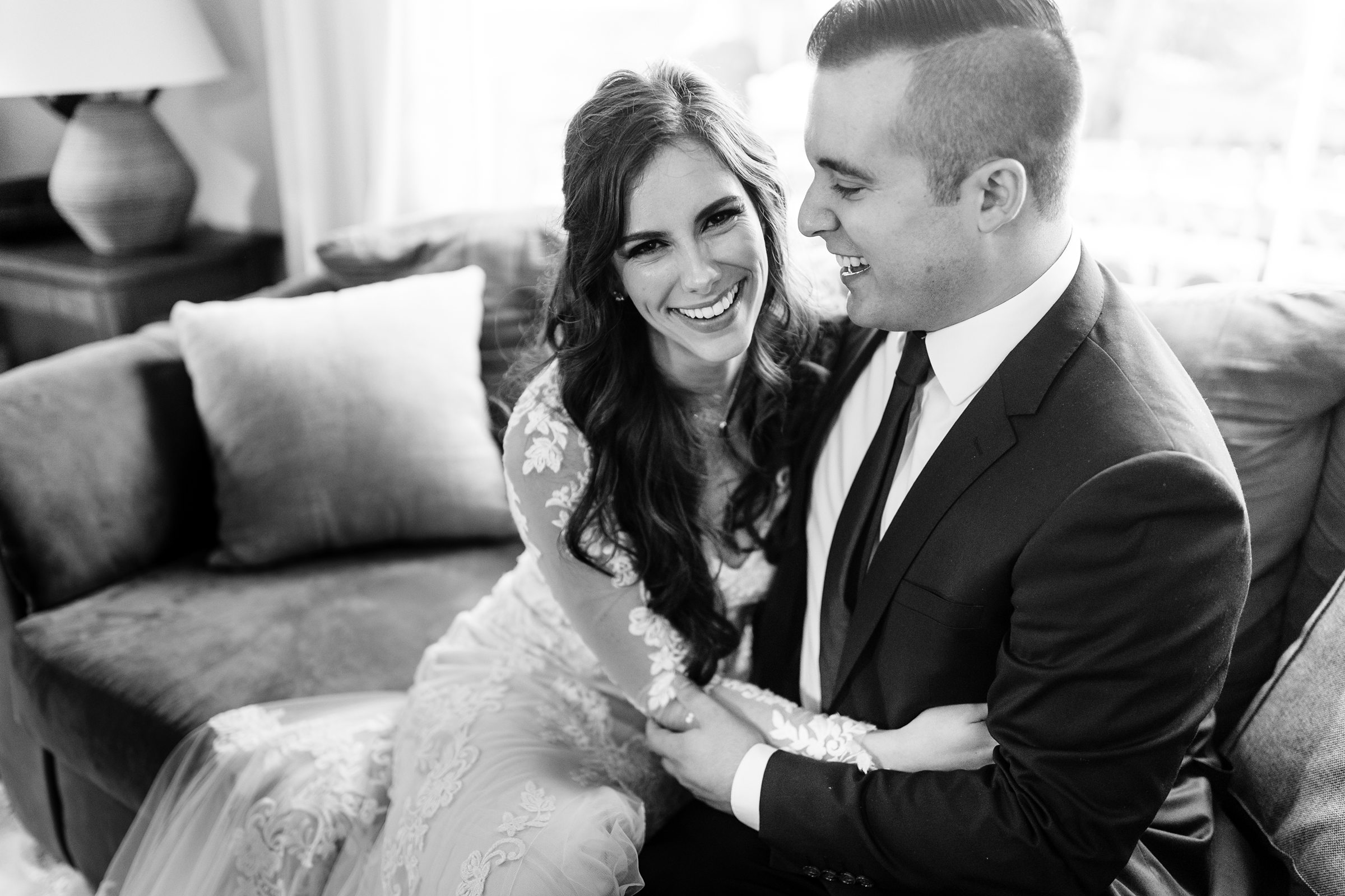 Black and white photo of bride and groom sitting on couch and cuddling, bride is smiling for camera and groom is looking at bride by Detroit Wedding Photographer Michele Maloney