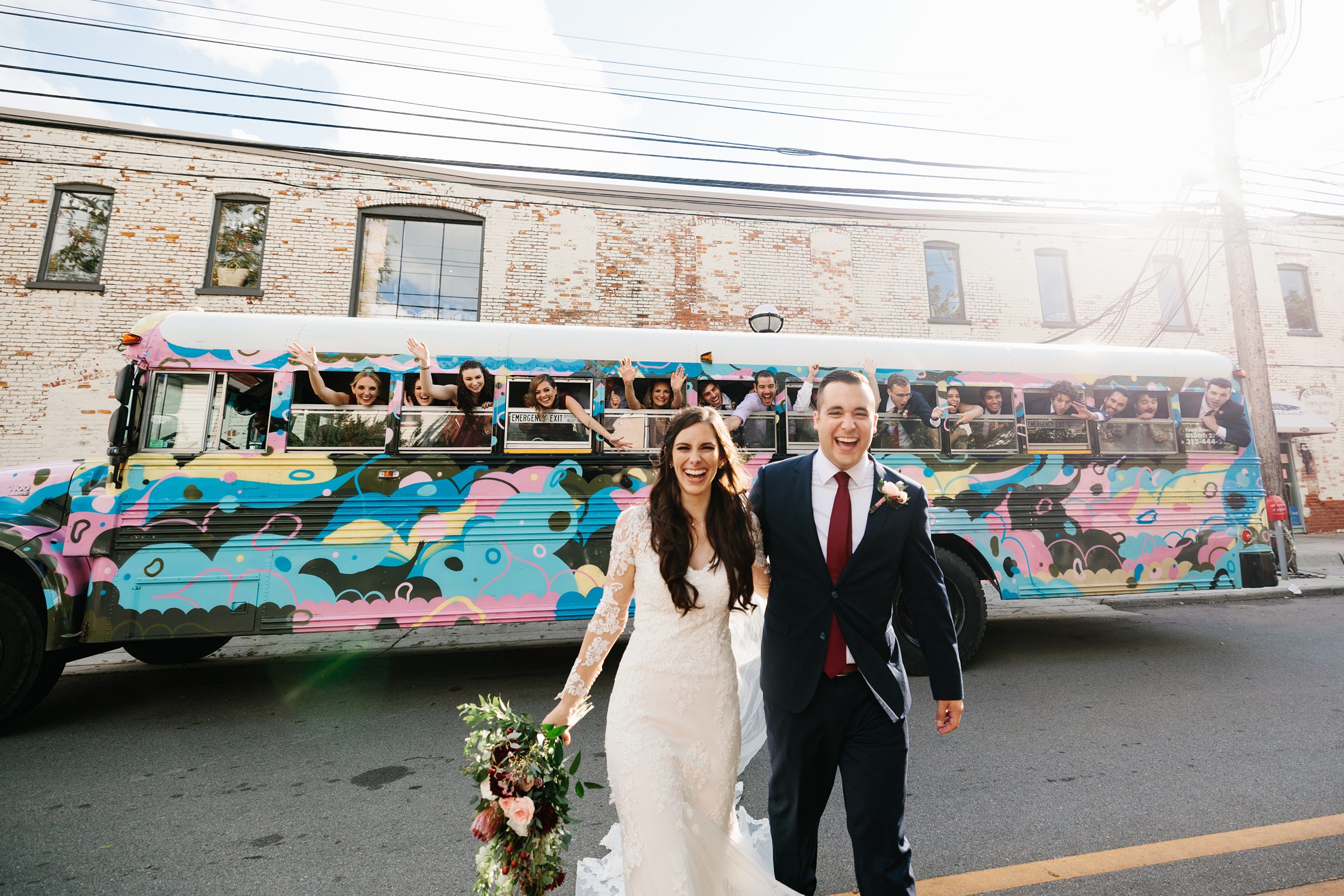 Bride and groom stand together and smile with the party bus behind them full of the bridal party smiling and waving at the camera for the Ann Arbor intimate wedding by Detroit Wedding Photographer Michele Maloney 