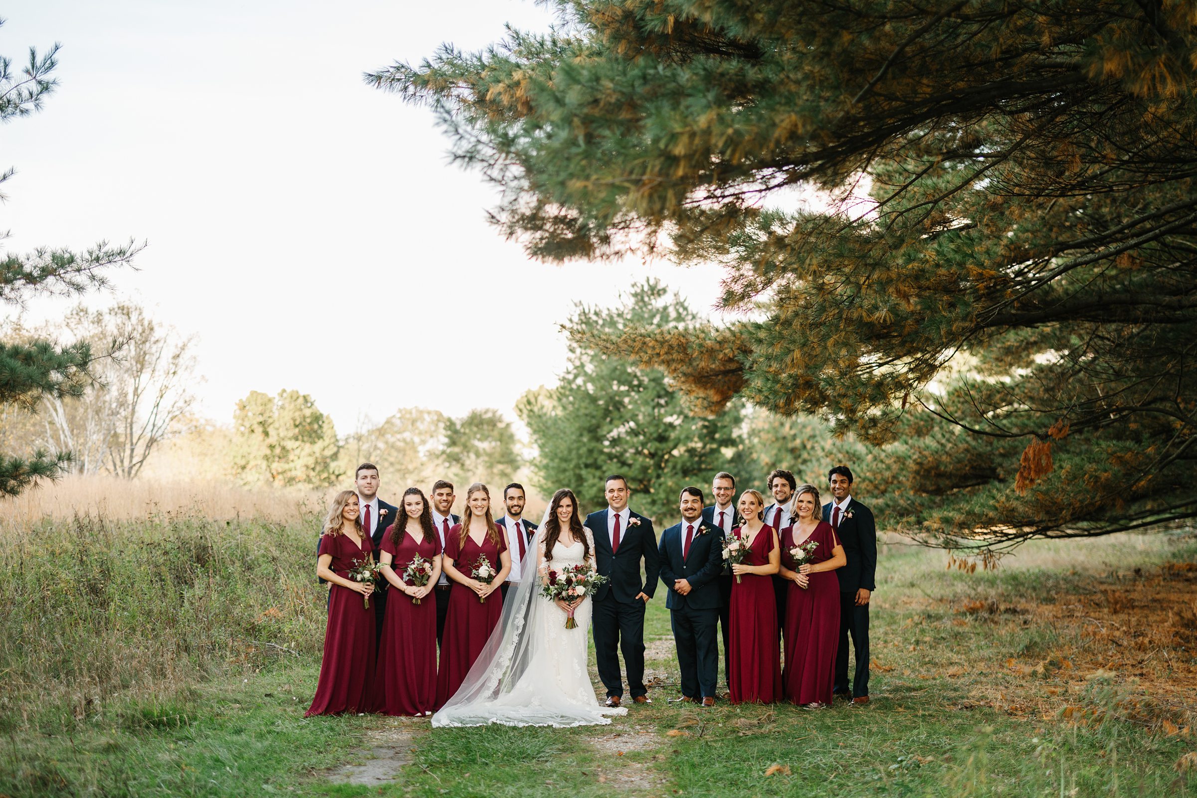 Bride and groom are surrounded by their bridal party underneath a pine tree, all are smiling at the camera for the Ann Arbor intimate wedding by Detroit Wedding Photographer Michele Maloney