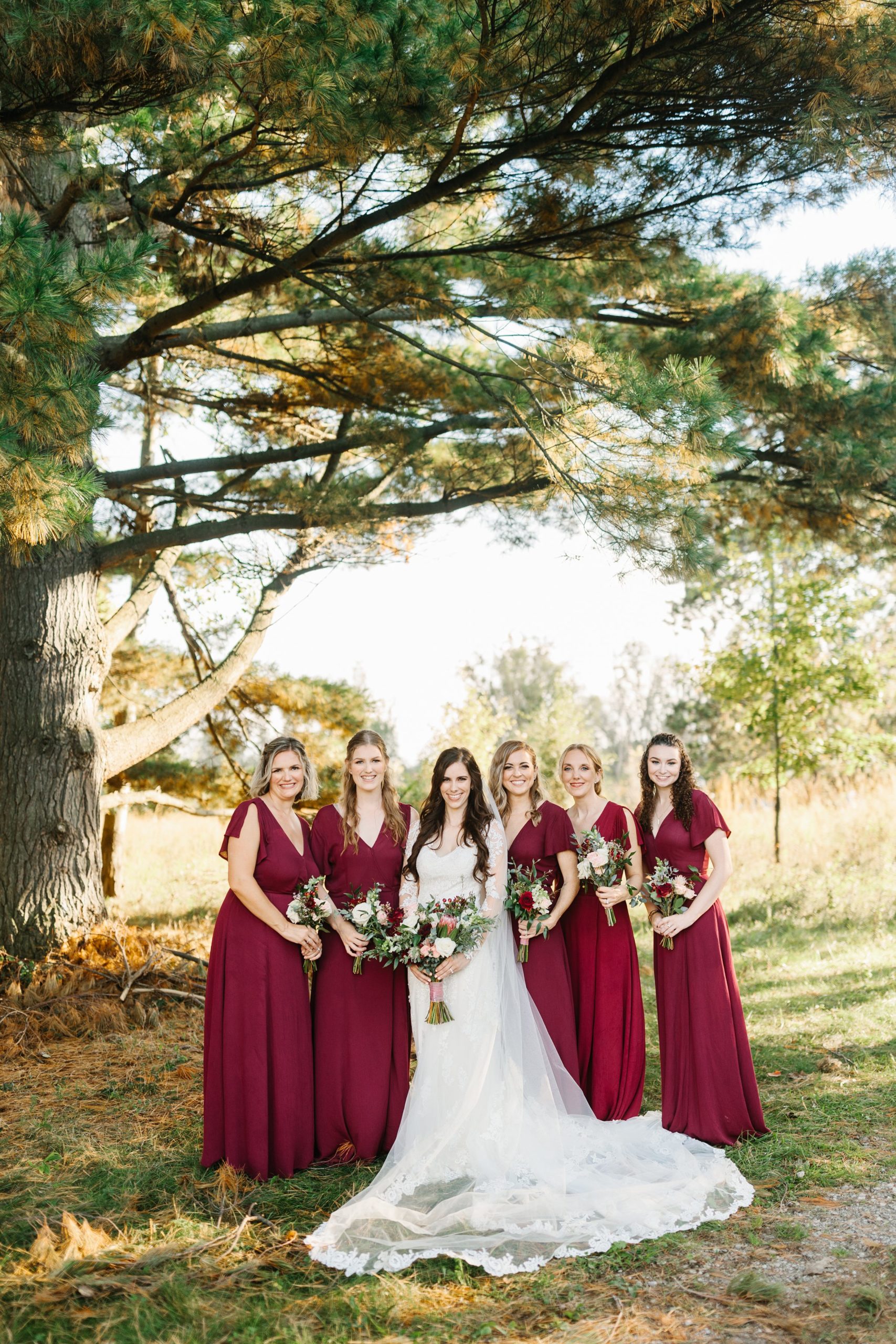 Bride stands in the middle with her bridesmaids around her, all are smiling for a portrait under a pine tree by Detroit Wedding Photographer Michele Maloney