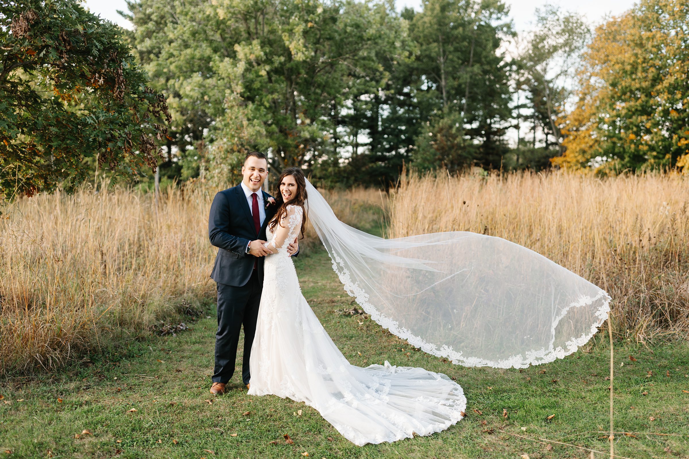 Bride and groom smile for the camera and hold each other, brides long veil billows in the wind by Detroit Wedding Photographer Michele Maloney