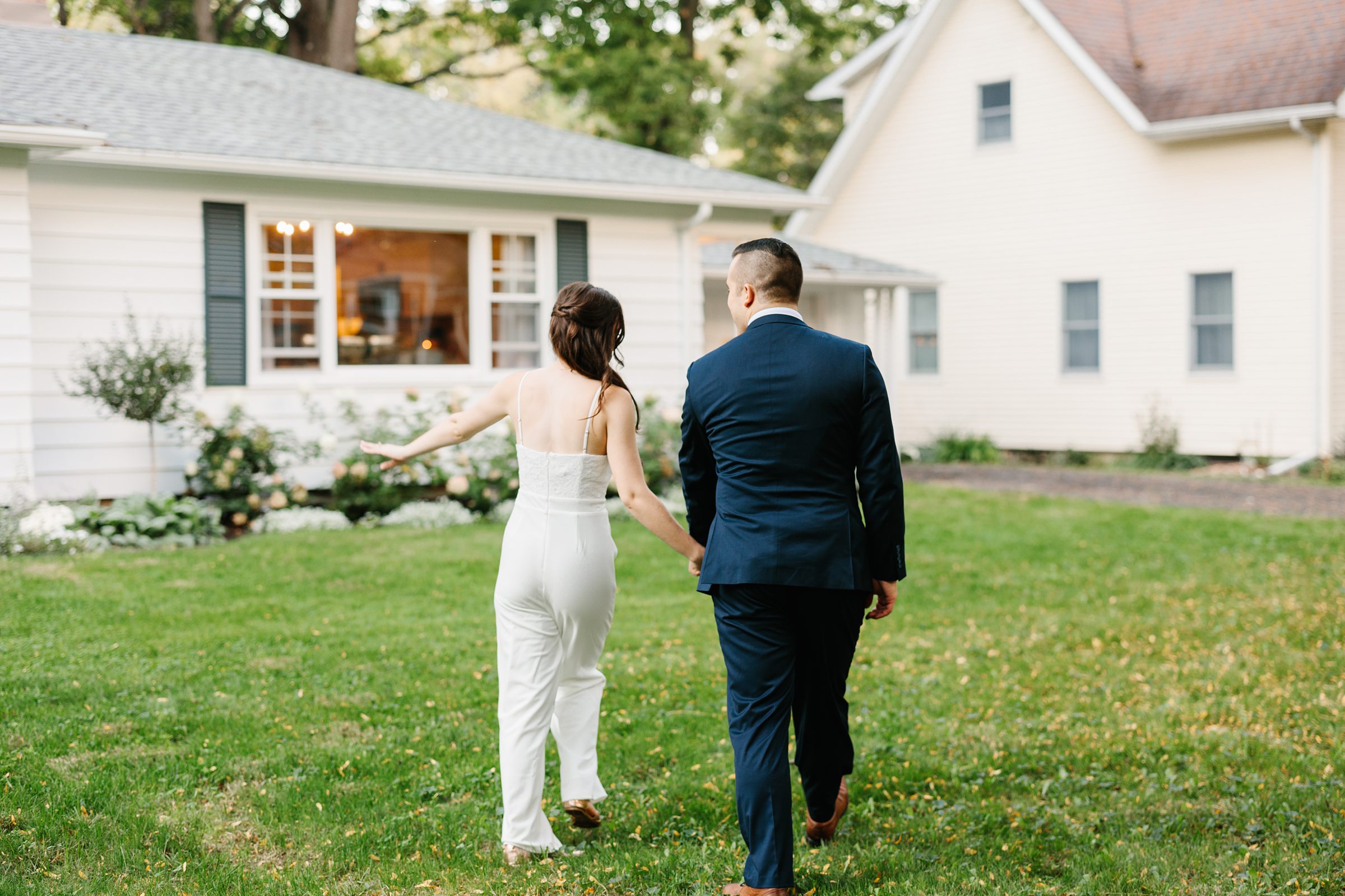 Shot from behind bride and groom holding hands while walking in the backyard of their Ann Arbor intimate wedding by Detroit Wedding Photographer Michele Maloney