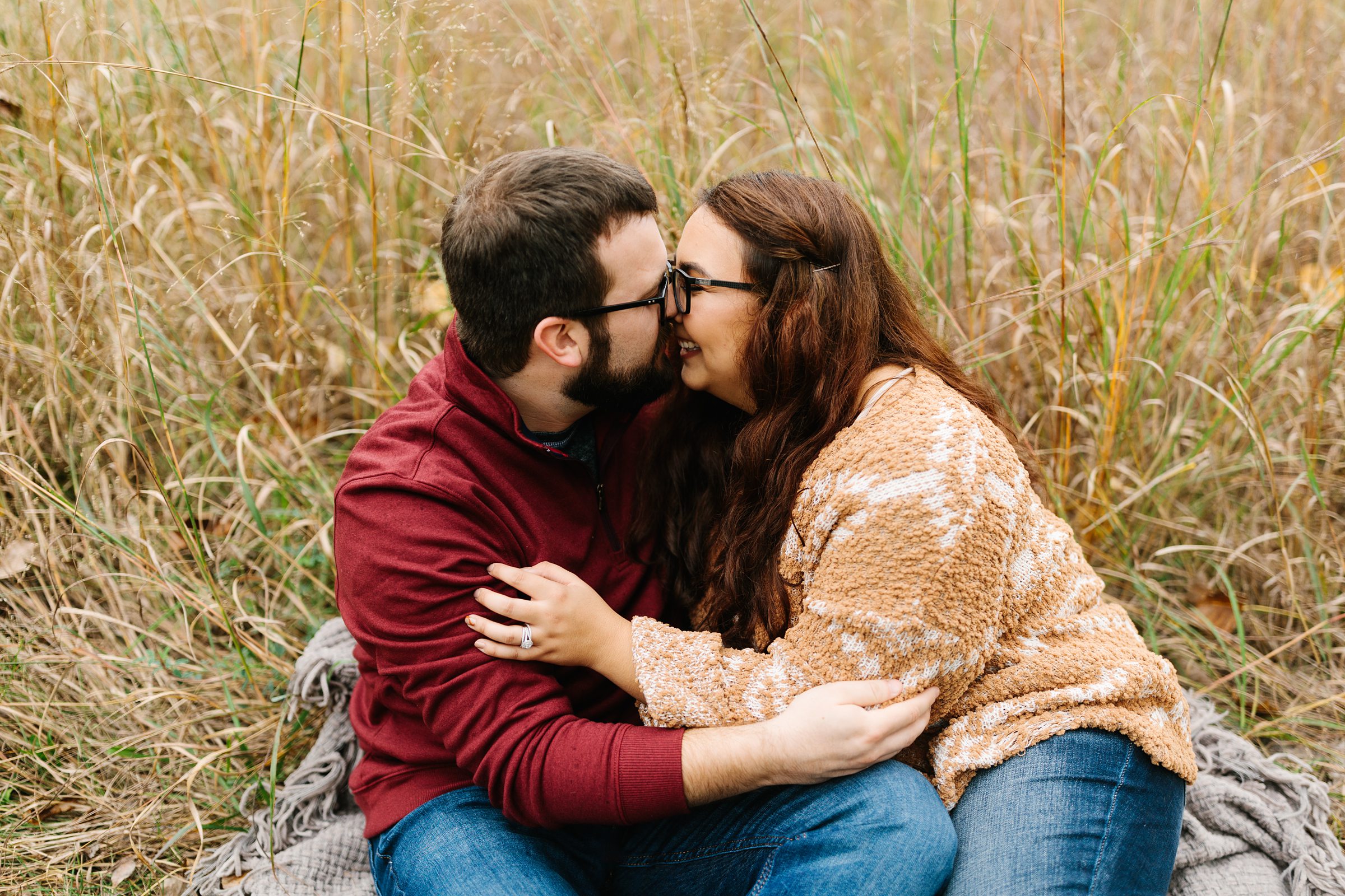 Engaged couple sits outside and cozies up close, giving eskimo kisses in a grassy patch by Detroit Wedding Photographer Michele Maloney