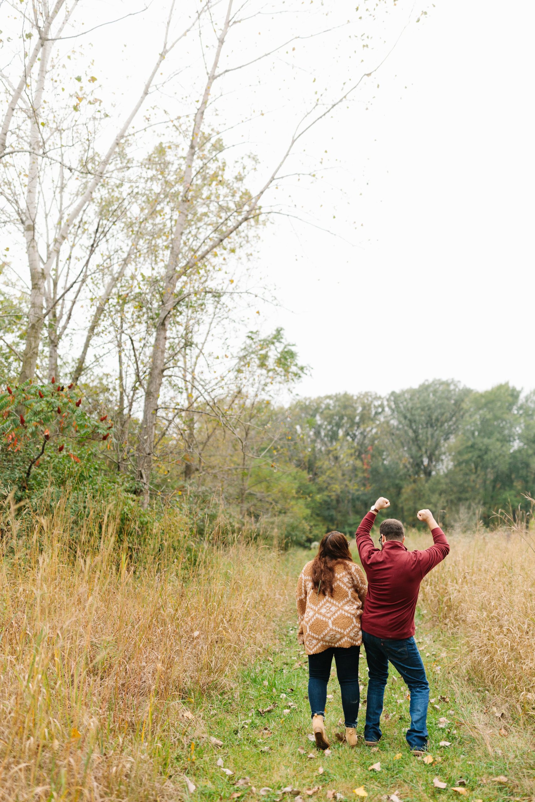Faraway shot of engaged couple from behind walking in a grassy path, fiancé lifts up his hands and playfully bumps his future bride by Detroit Wedding Photographer Michele Maloney 