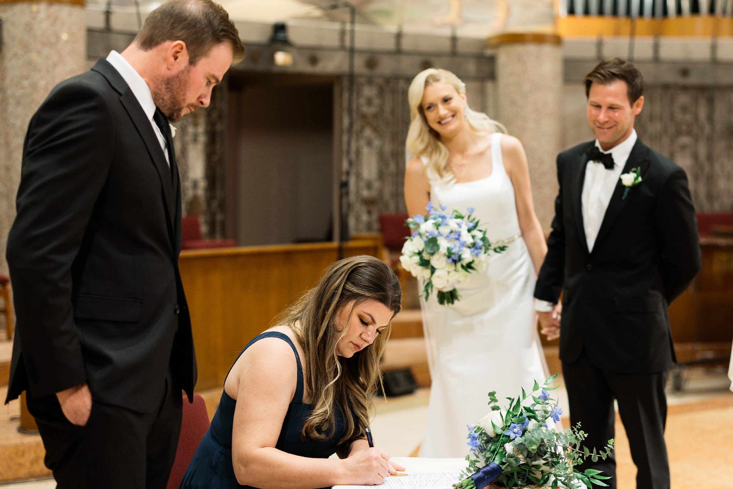 Bridesmaid and groomsmen sign as witnesses to marriage certificate, bride and groom hold hands and watch by Detroit Wedding Photographer Michele Maloney 