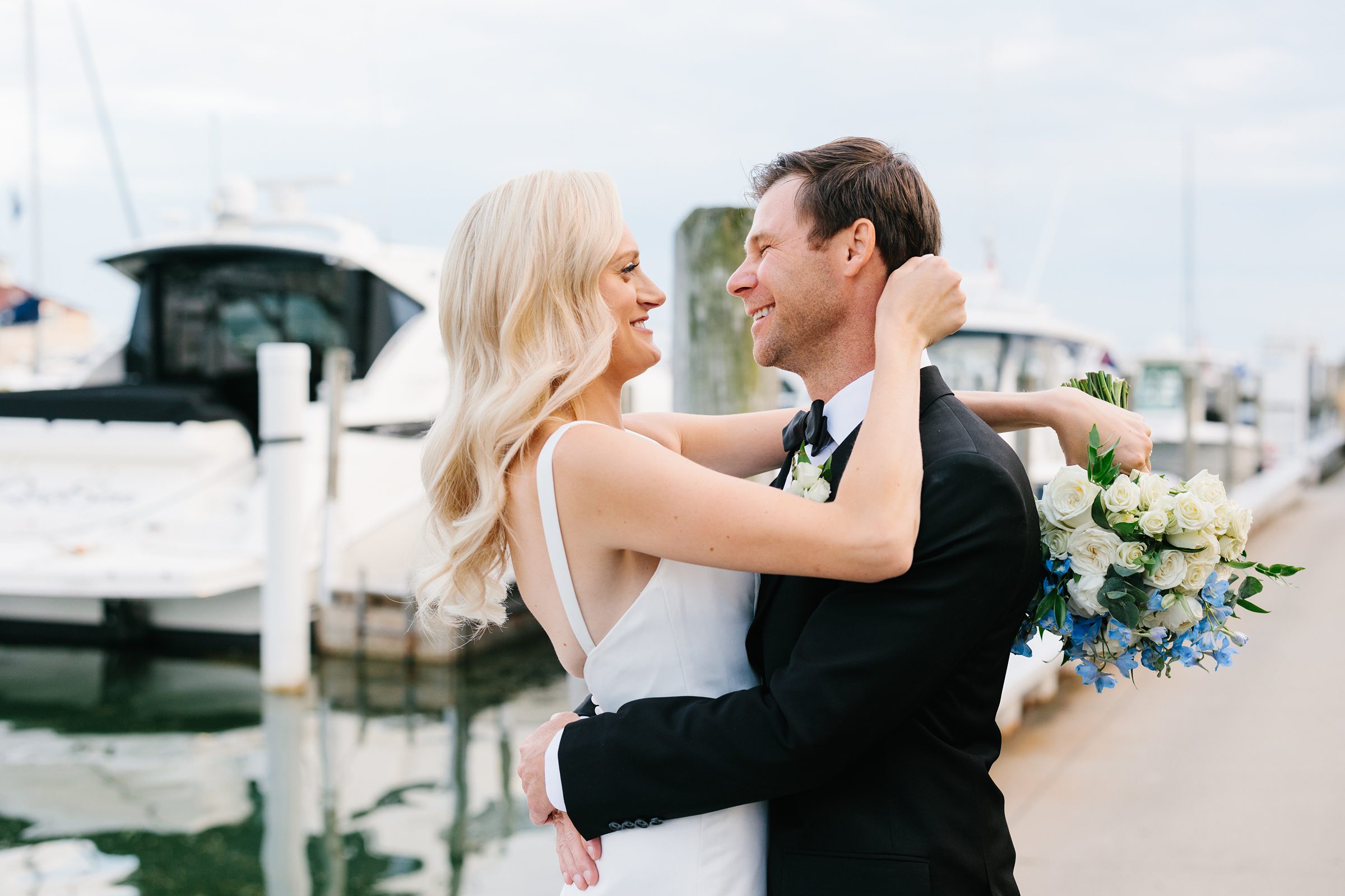 Bride hugs groom around his neck and they smile near the dock at the Grosse Pointe Yacht Club by Detroit Wedding Photographer Michele Maloney