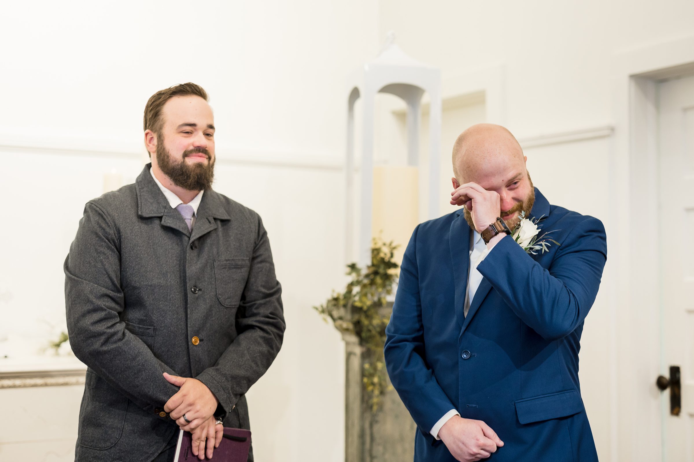 Officiant and groom watch bride come down the aisle, groom is wiping away his tears at the All Saints Wedding Chapel by Detroit Wedding Photographer Michele Maloney