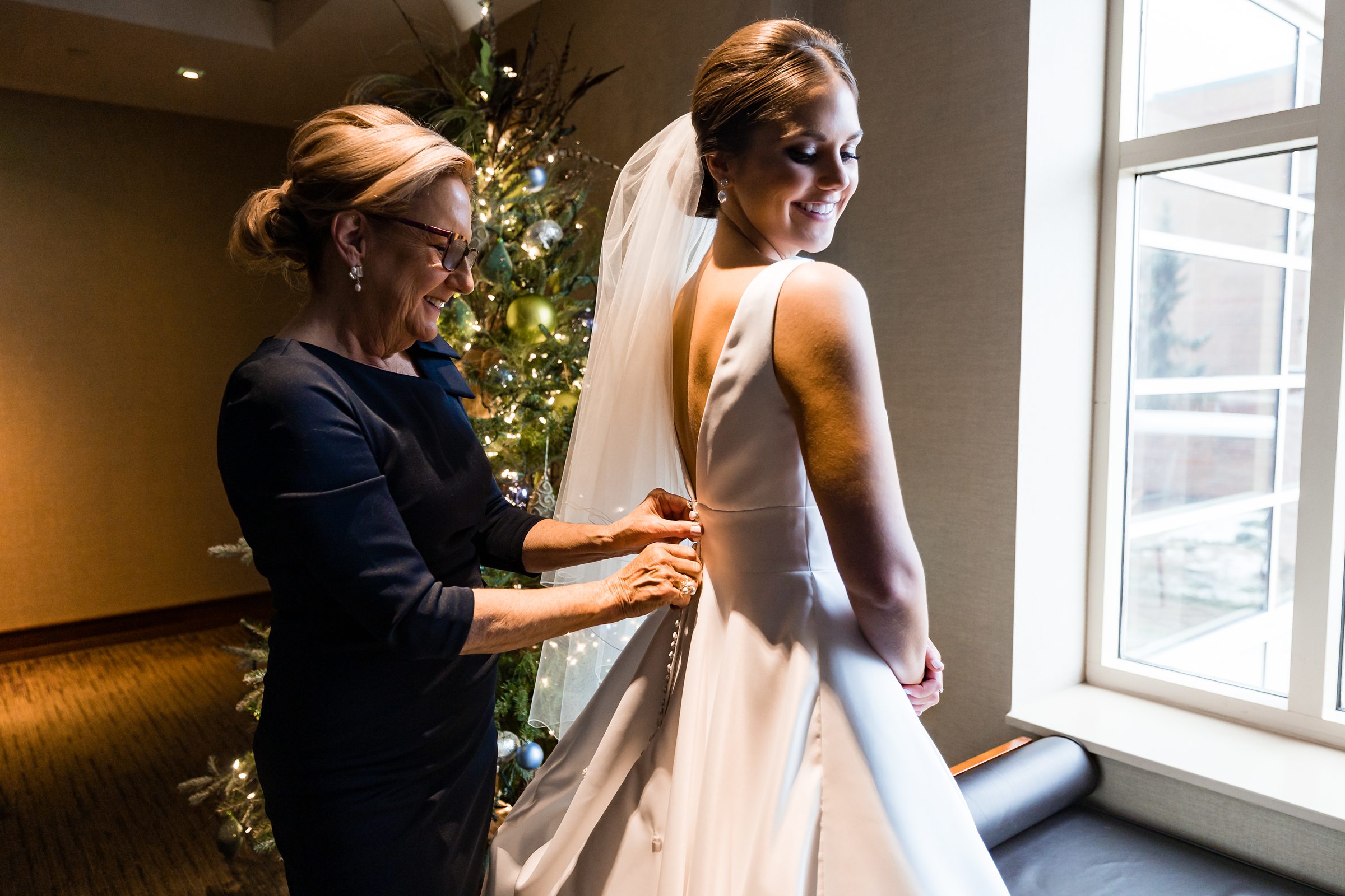 Mother of the bride buttons up the brides dress, both mother and daughter are smiling by Detroit Wedding Photographer Michele Maloney?