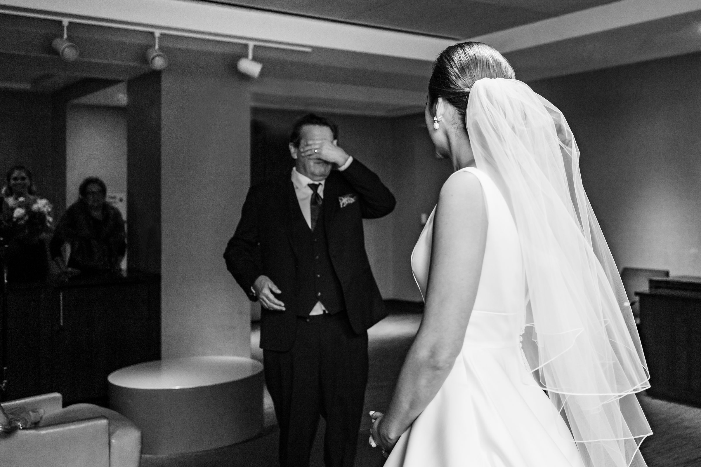 Black and white photo of father of the bride covering his eyes as bride enters so he can see her for the first time by Detroit Wedding Photographer Michele Maloney 