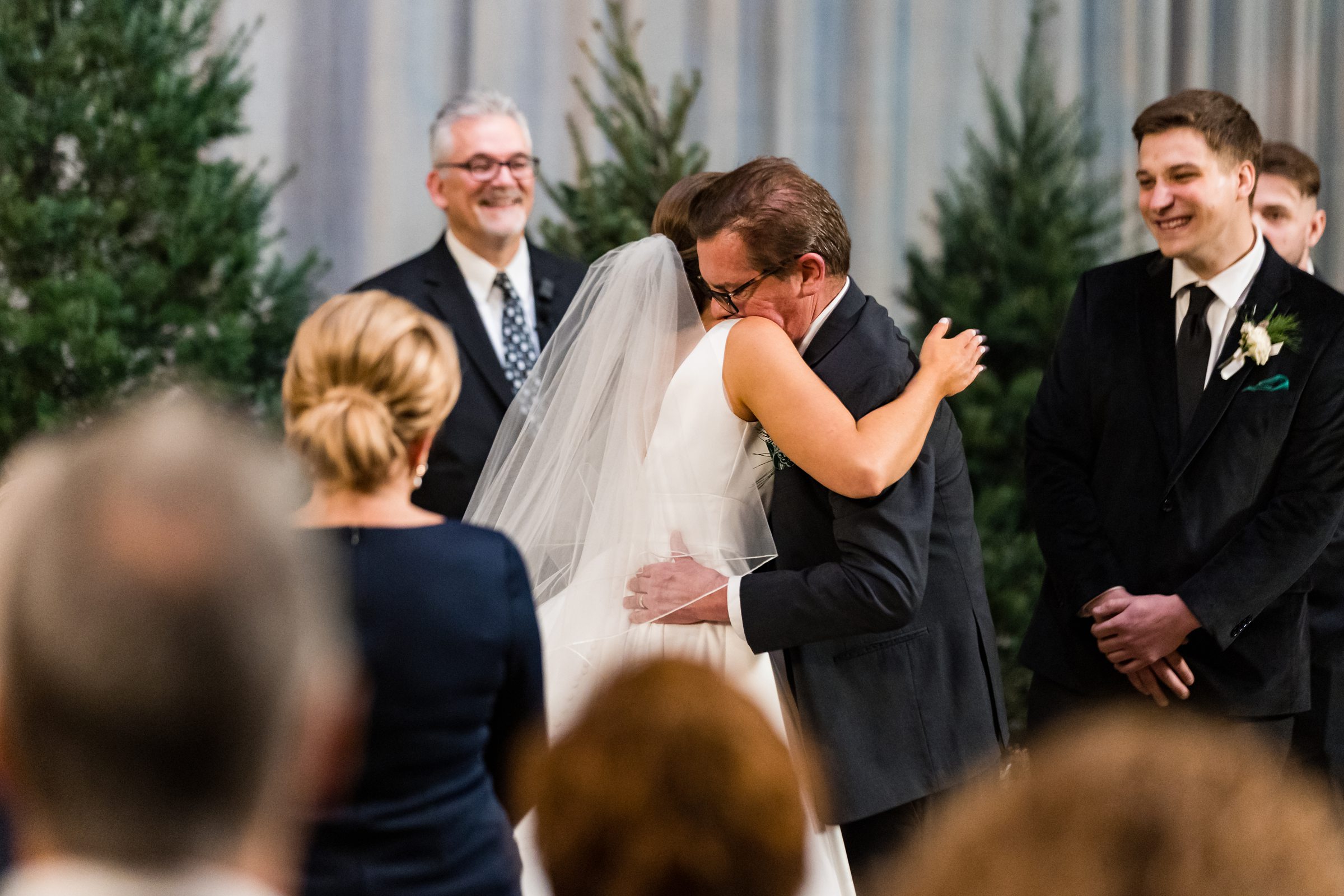 Bride and father of the bride hug at the end of the aisle, groom and mother watch and smile by Detroit Wedding Photographer Michele Maloney