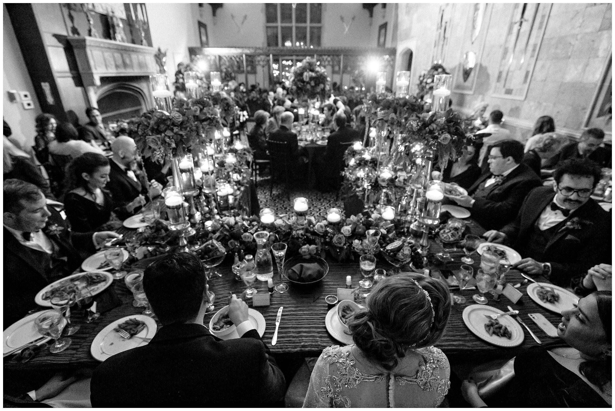 Black and white photo from behind the bride and groom sitting at the reception with all of their loved ones gathered around eating and celebrating by Detroit Wedding Photographer Michele Maloney