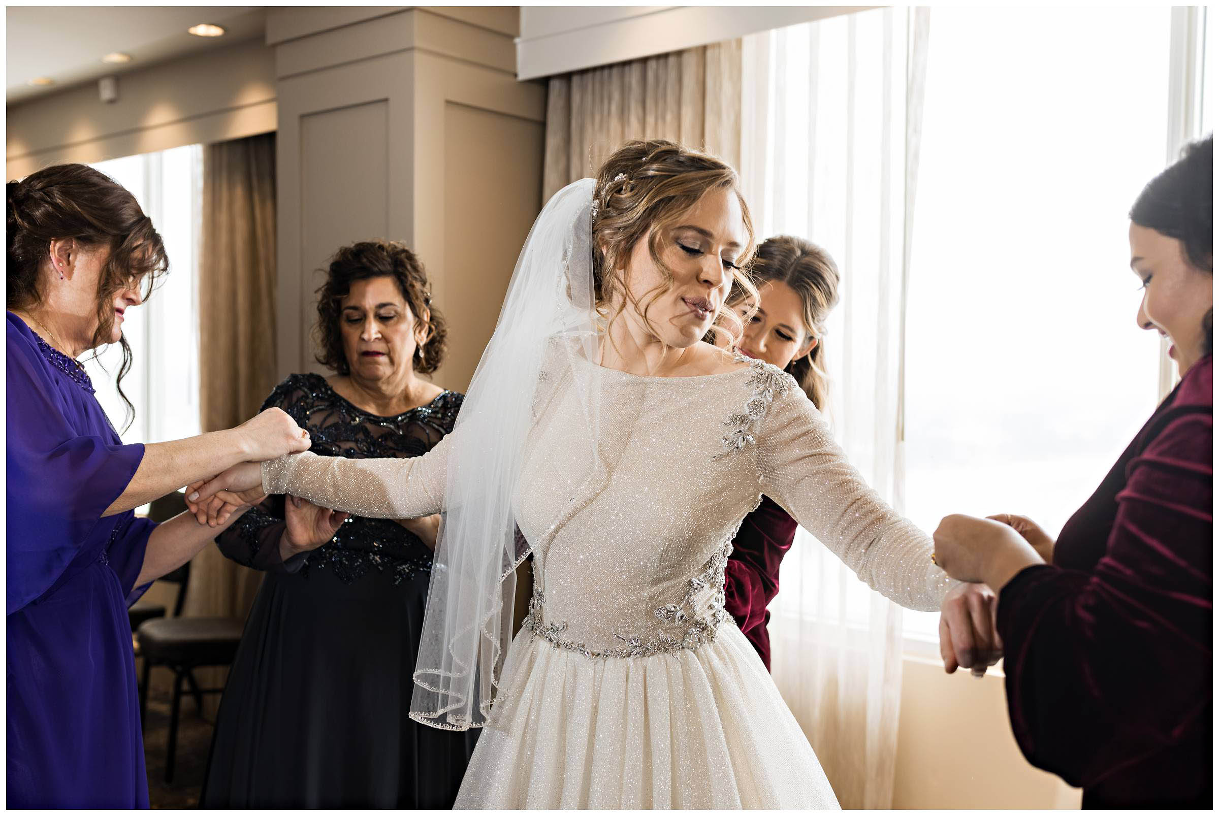 Bride standing in the center as her friends and family adjust her wedding dress by Detroit Wedding Photographer Michele Maloney