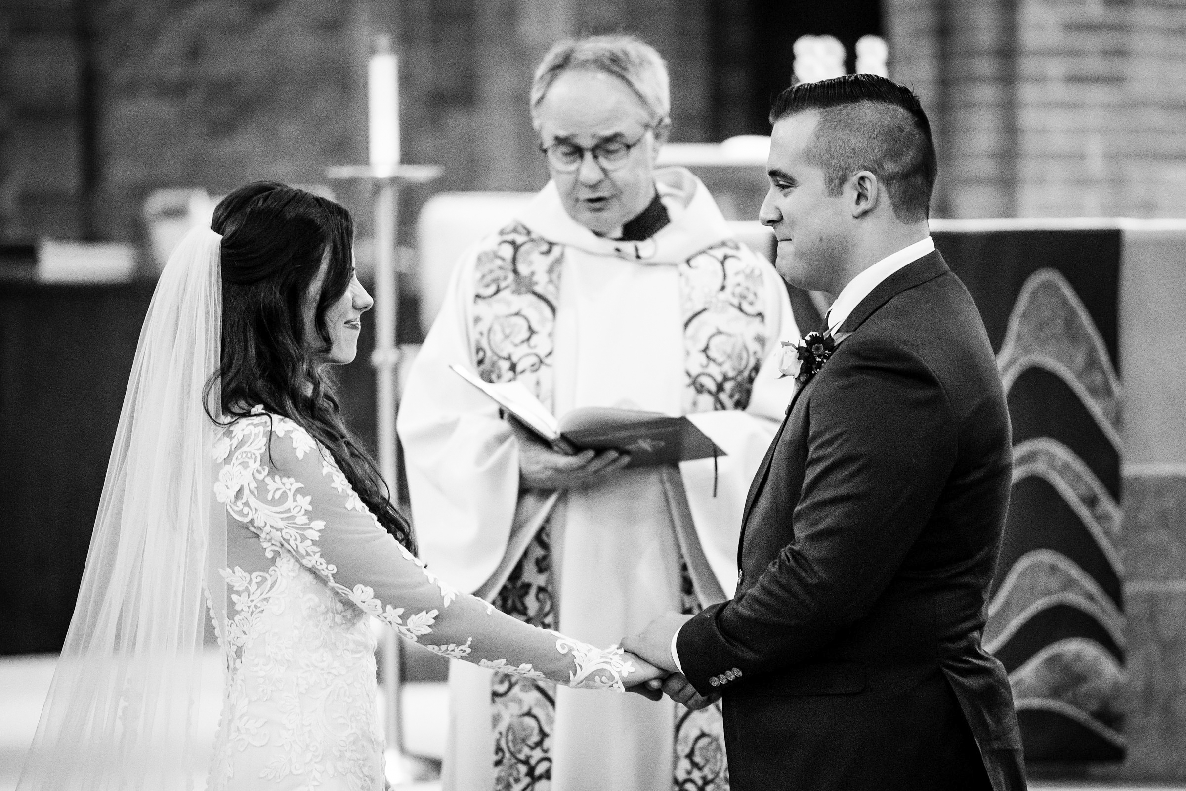 Black and white photo of bride and groom holding hands as they face each other on stage for their ceremony with the priest officiating in the background by Detroit Wedding Photographer Michele Maloney 