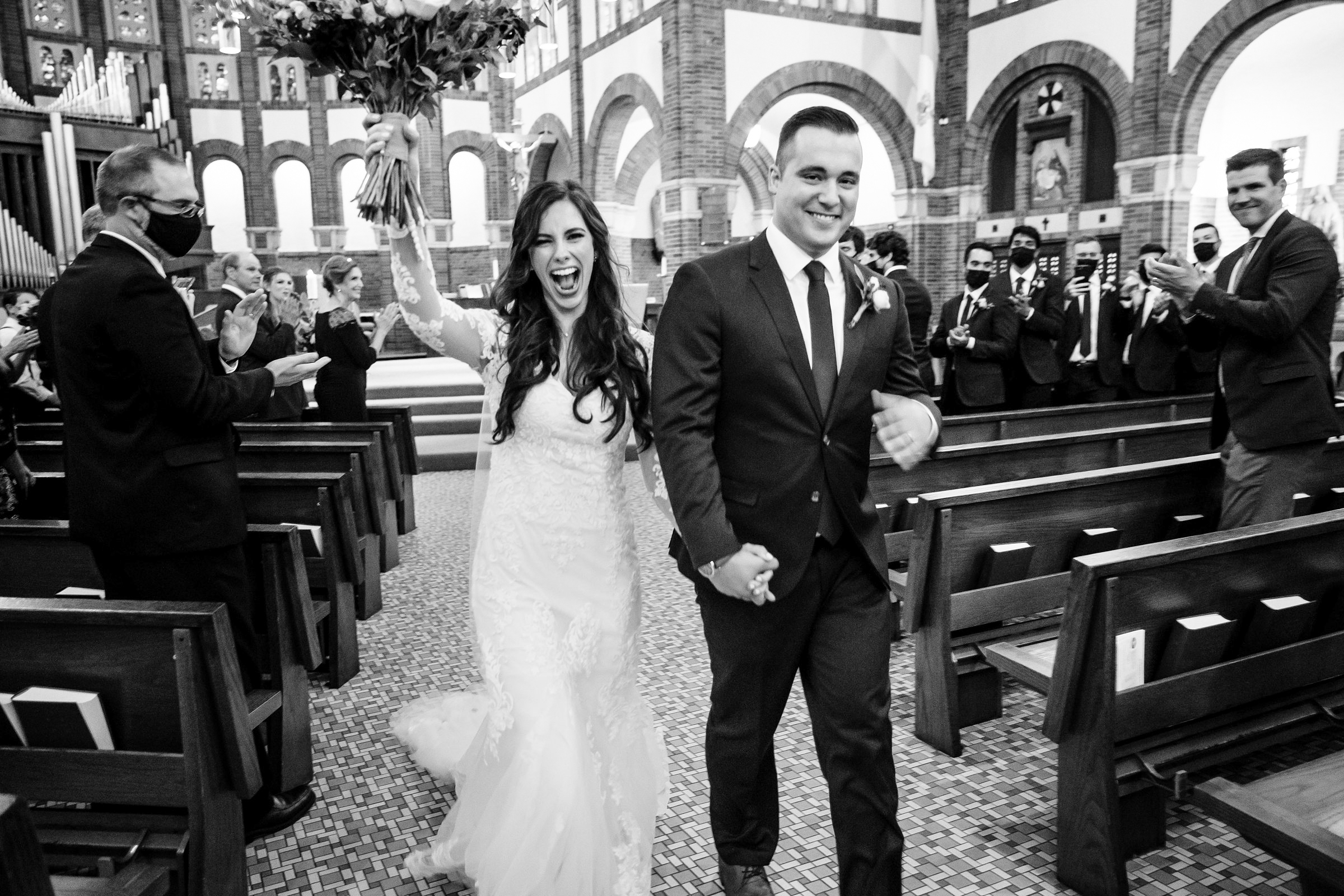 Black and white photo of bride and groom reaching the end of the aisle for their recessional, bride holds up her bouquet and they're both grinning by Detroit Wedding Photographer Michele Maloney