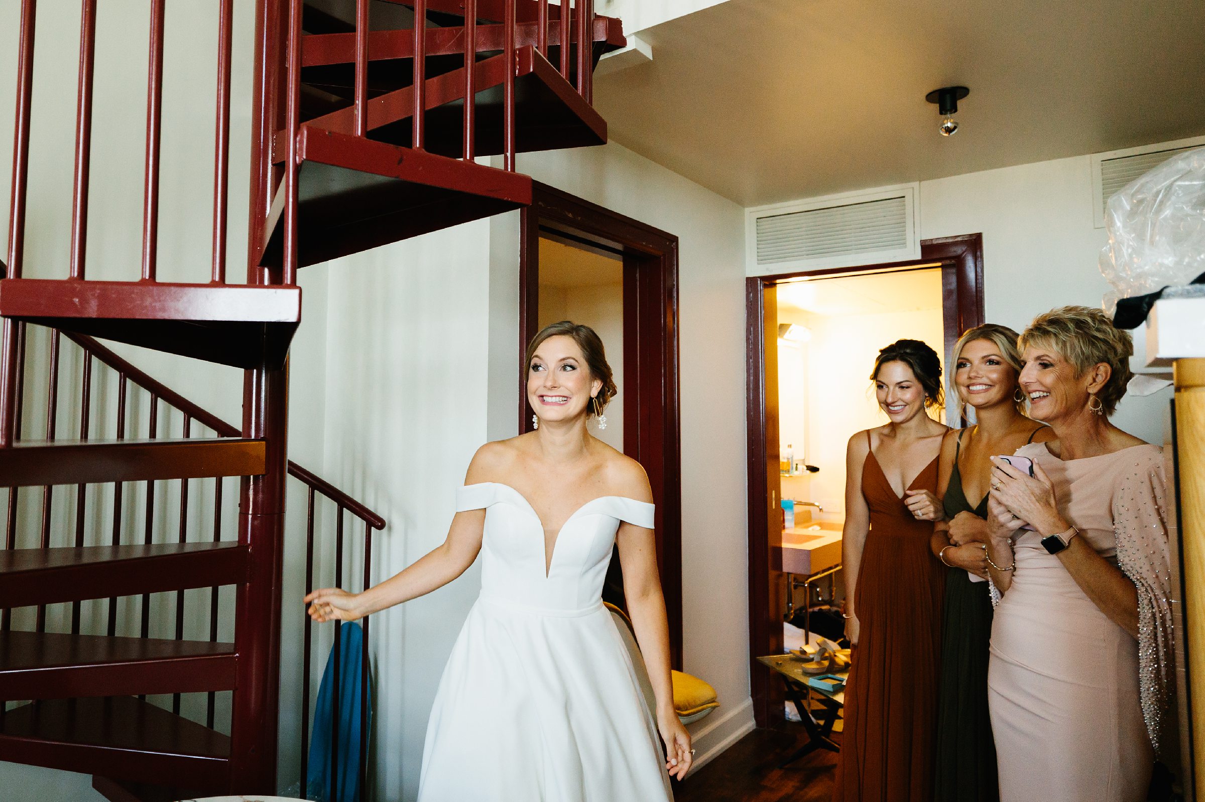 Bride grins ear to ear beside the spiral staircase in the Siren Hotel and the bridesmaids and mother of the bride smile at the bride by Detroit Wedding Photographer Michele Maloney 