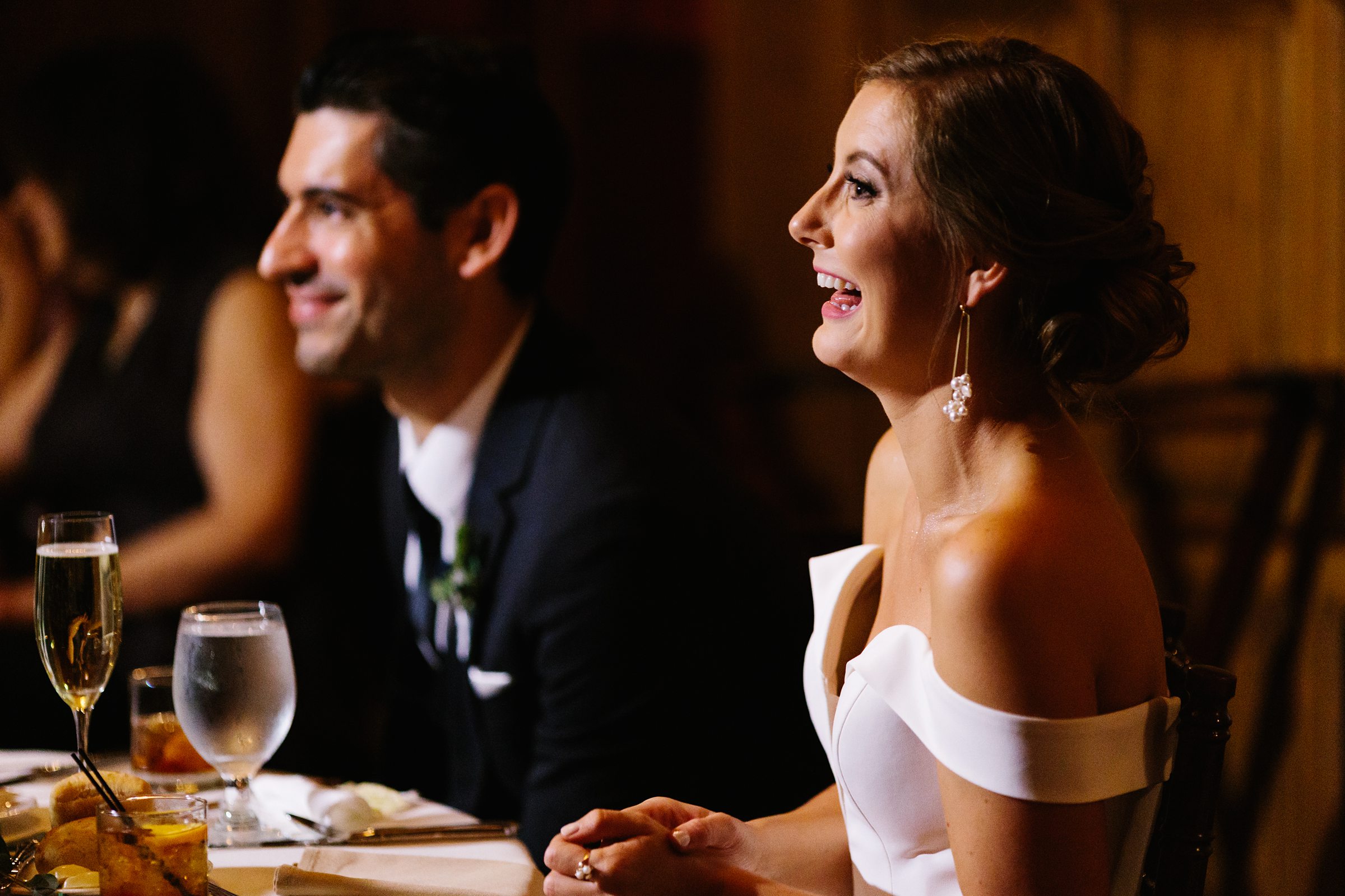 Bride and groom smile and laugh as they sit and listen to toasts at the Gem Theater wedding by Detroit Wedding Photographer Michele Maloney