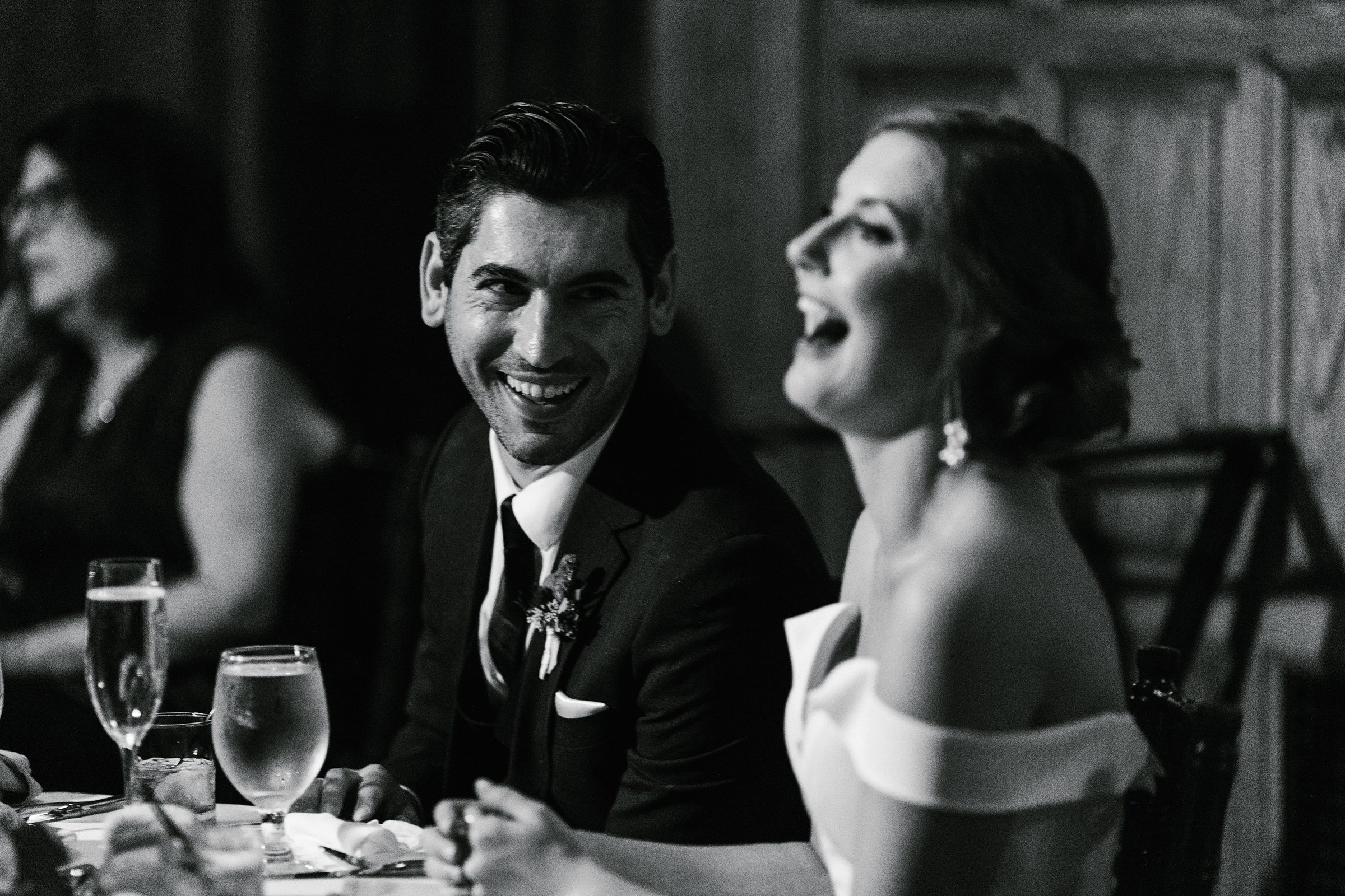 Black and white photo of the groom watching his new bride laugh while she listens to wedding toasts by Detroit Wedding Photographer Michele Maloney