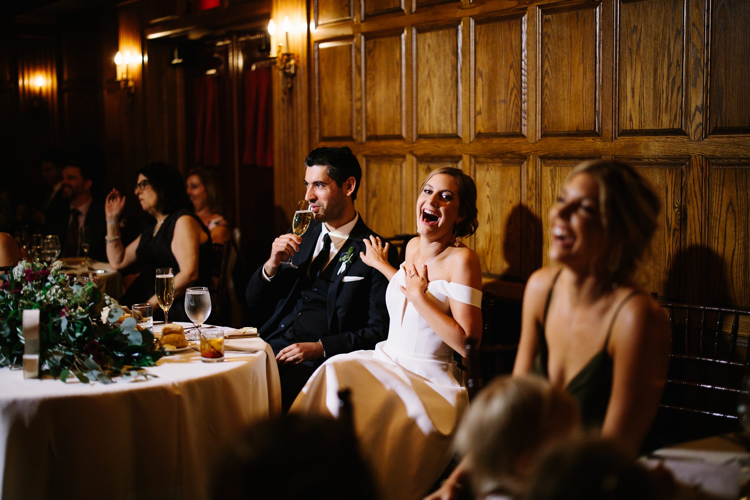 Bride laughs and touches groom as he sips his drink during wedding toasts by Detroit Wedding Photographer Michele Maloney