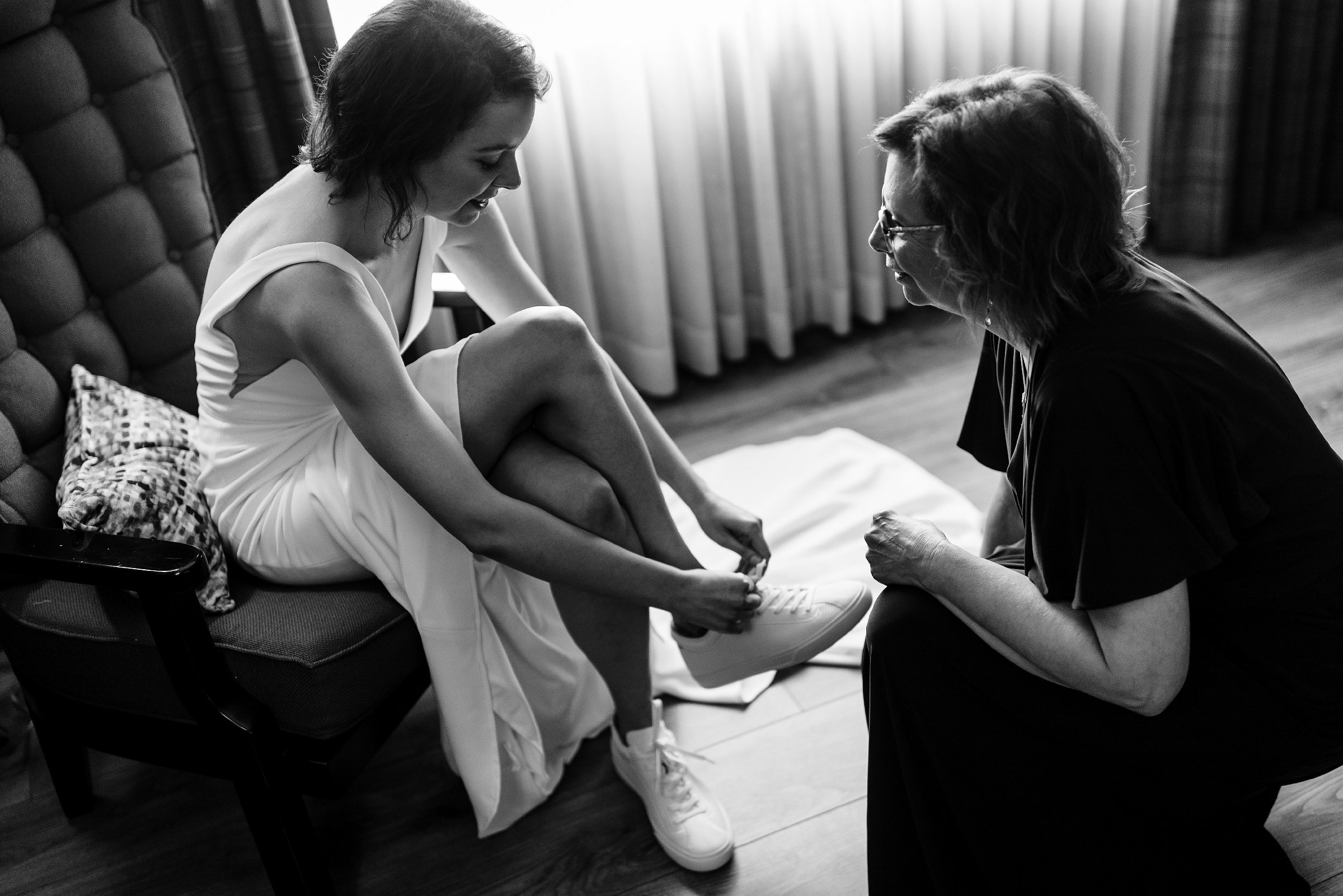 Black and white picture of the bride sitting on a share and tying her shoes, mother of the bride sits next to her and looks at her by Detroit Wedding Photographer Michele Maloney