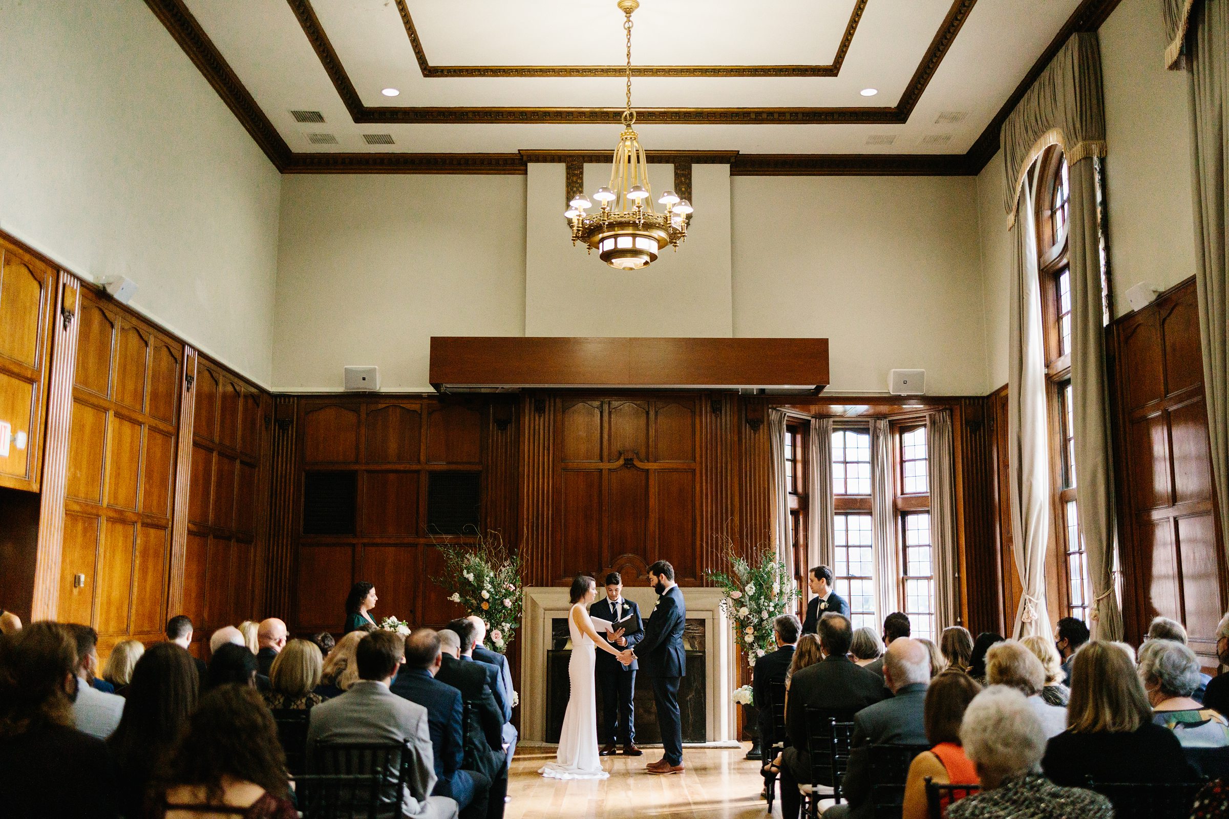 Faraway shot of the bride and groom holding hands and looking at each other during the ceremony with guests all watching by Detroit Wedding Photographer Michele Maloney