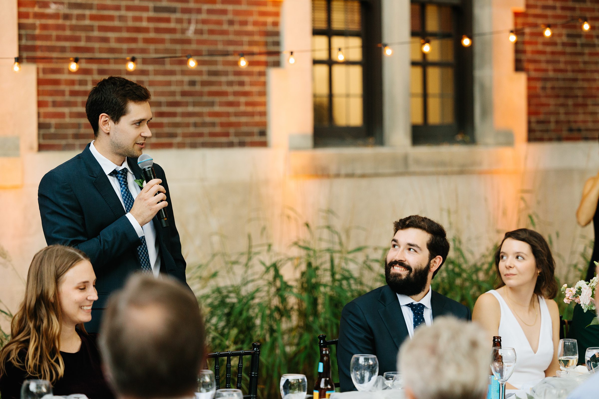 Groomsmen holds mic and gives his toast while bride and groom smile during their outdoor wedding reception under cafe lights by Detroit Wedding Photographer Michele Maloney