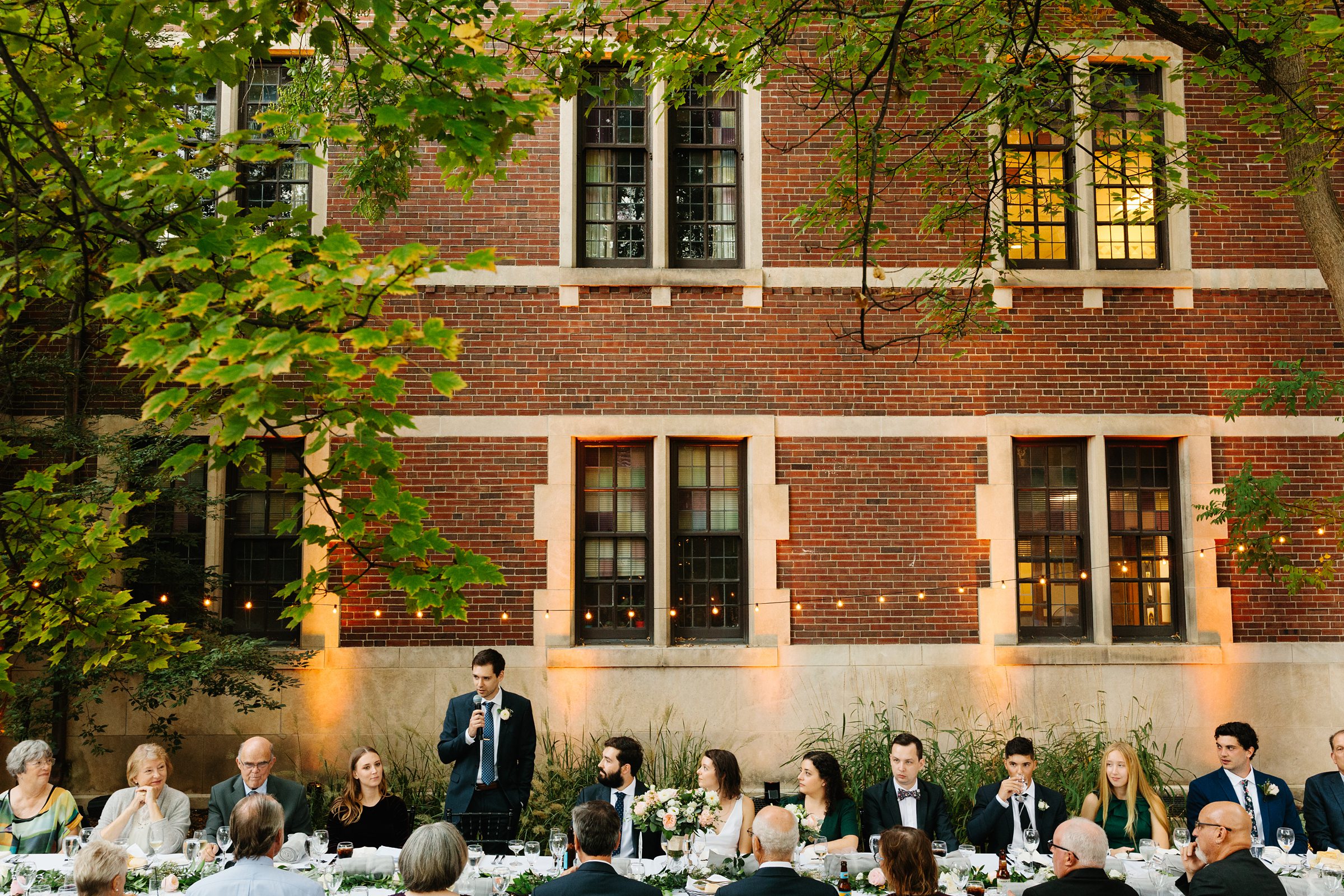 Faraway shot of a wedding toast outdoors, guests all watch the groomsmen as he holds the mic by Detroit Wedding Photographer Michele Maloney