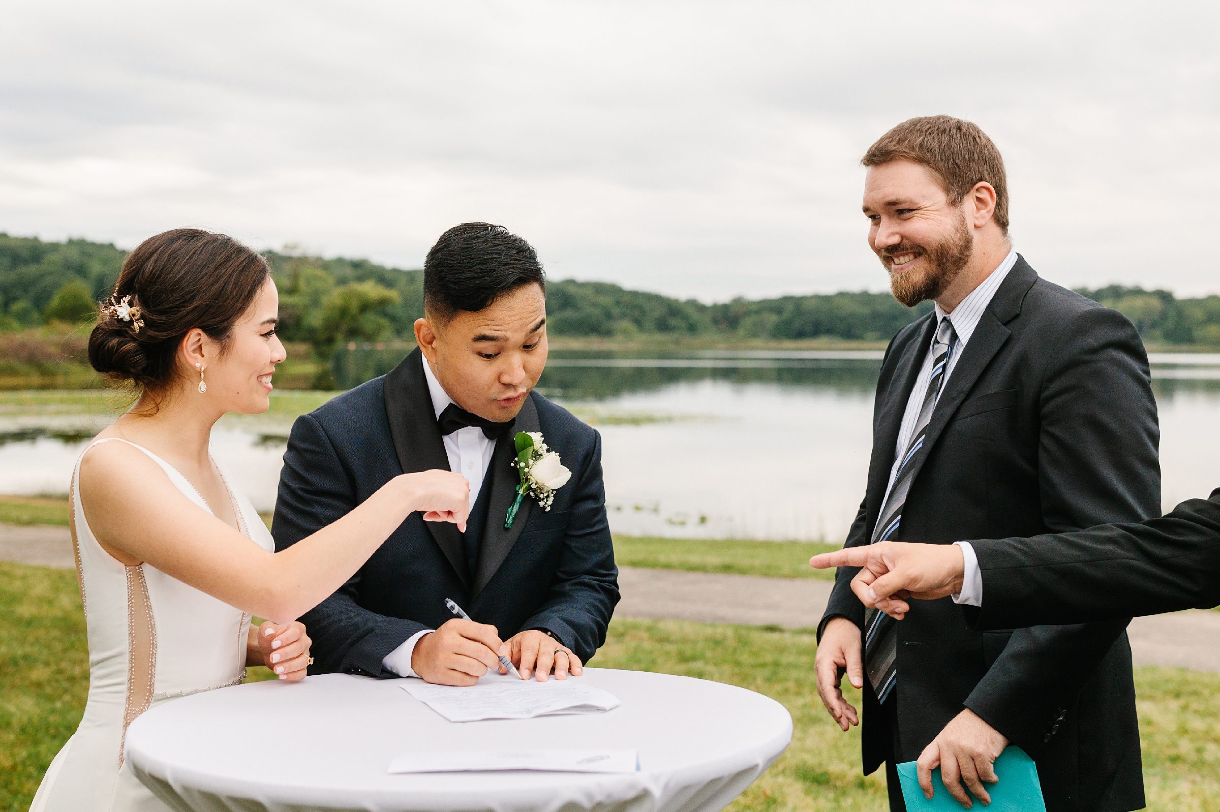 Pastor and groom are talking while they sign marriage certificate, bride is smiling by Detroit Wedding Photographer Michele Maloney