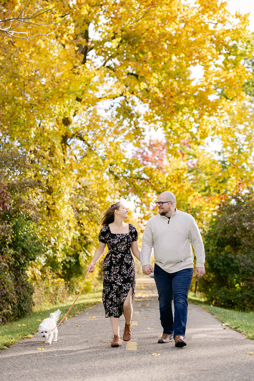 Brooke and Steven walking along with their pup on a leaf-covered path at Lake Erie Metropark during fall.